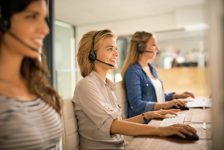 Three female customer service representatives with headsets, reviewing claims on computer screens in an office setting